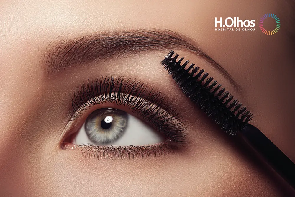 Close-up do olho claro de uma mulher branca maquiada, com uma escova próxima à sobrancelha, ilustrando cuidados com Maquiagem e lente de contato. No canto superior direito, aparece o logotipo "H.Olhos Hospital de Olhos".