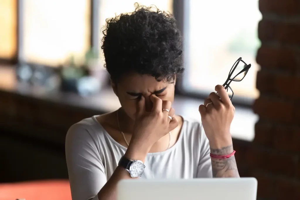 Mulher negra, de cabelo cacheado preso, aperta a região dos olhos com expressão de desconforto enquanto segura um par de óculos, em ambiente interno desfocado, representando sintomas de infecção da pálpebra.