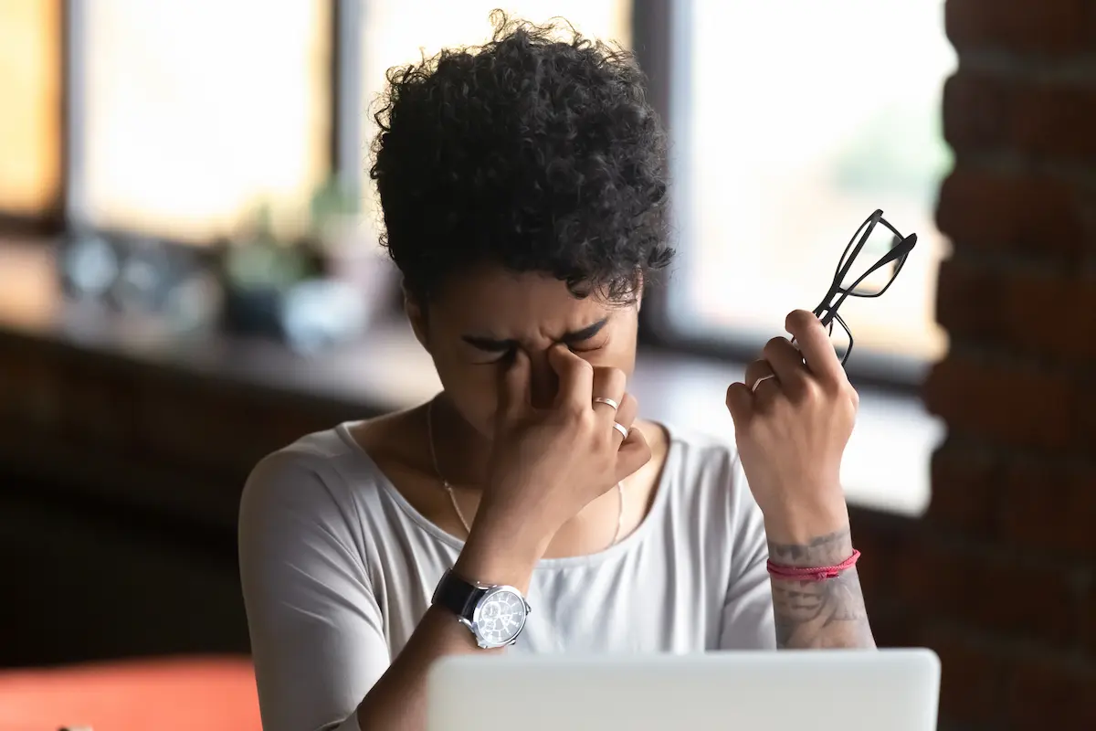 Mulher negra, de cabelo cacheado preso, aperta a região dos olhos com expressão de desconforto enquanto segura um par de óculos, em ambiente interno desfocado, representando sintomas de infecção da pálpebra.