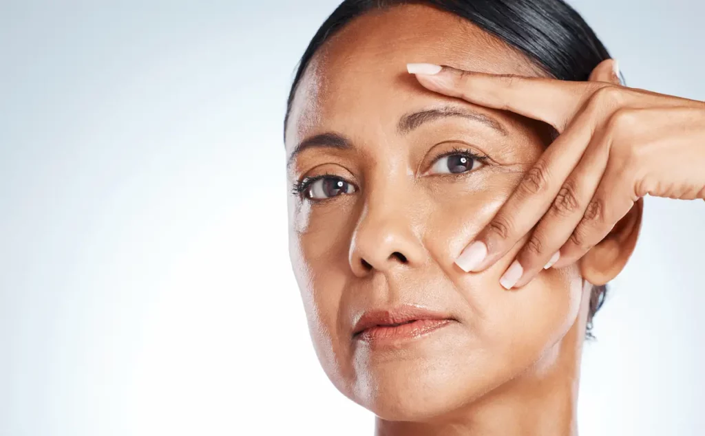 Mulher de meia-idade observa a câmera enquanto estica suavemente a pele ao redor de um dos olhos, destacando marcas de expressão.