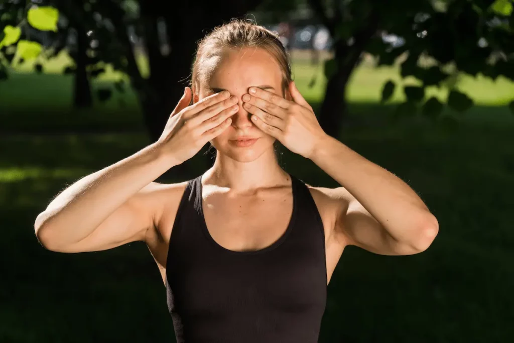 Mulher branca pratica palming ao ar livre, cobrindo os olhos fechados com as palmas das mãos em um parque arborizado.