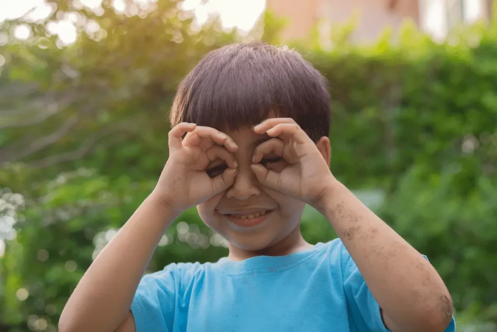 Menino asiático de camiseta azul leva as mãos aos olhos enquanto brinca ao ar livre, ilustrando a dúvida: O que devo fazer se meu filho machucar o olho?