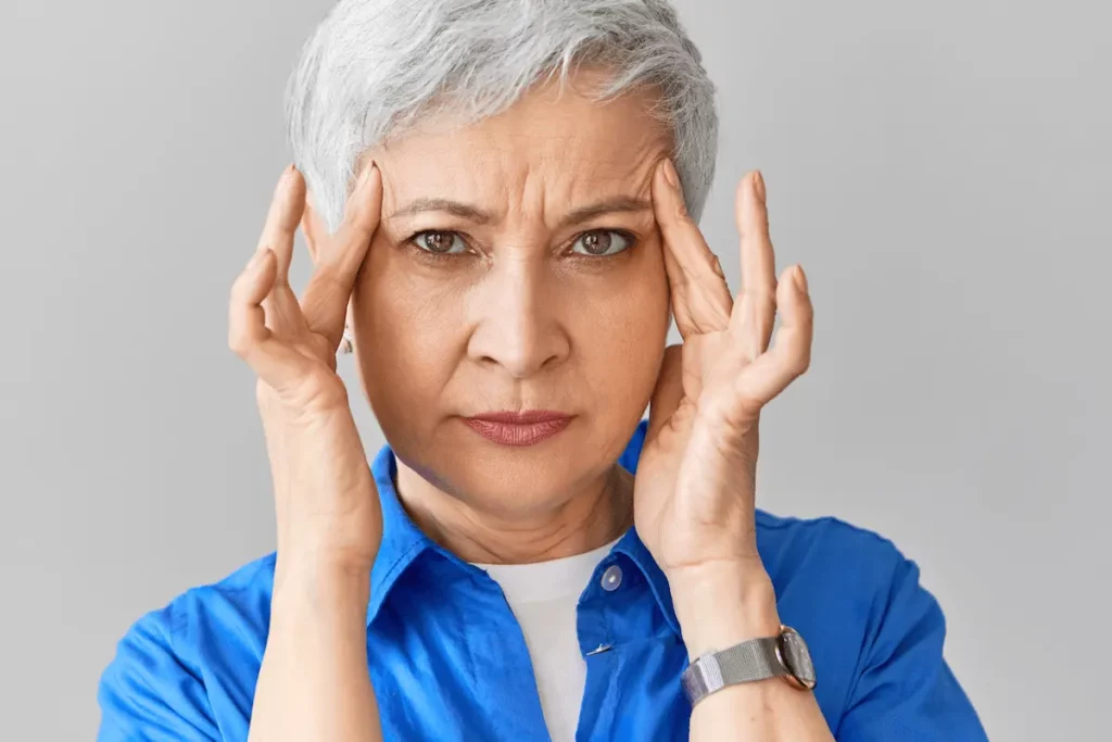Mulher branca de cabelos grisalhos pressiona as têmporas com expressão de incômodo, ilustrando a sensação de olhos pesados.