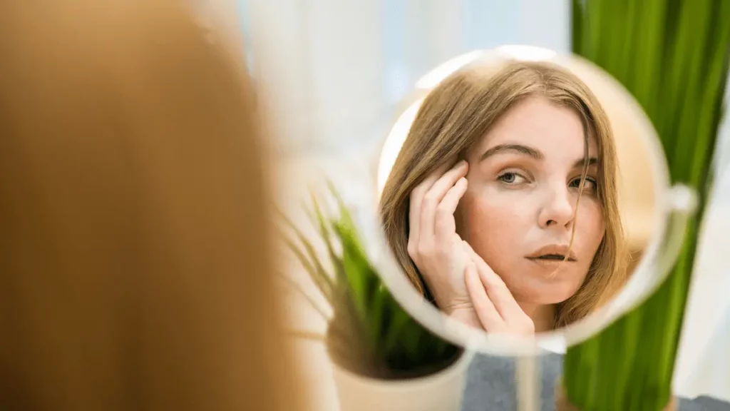 Mulher branca observa o próprio rosto no espelho enquanto toca a região abaixo dos olhos, em imagem relacionada à dúvida sobre por que olheiras aparecem.