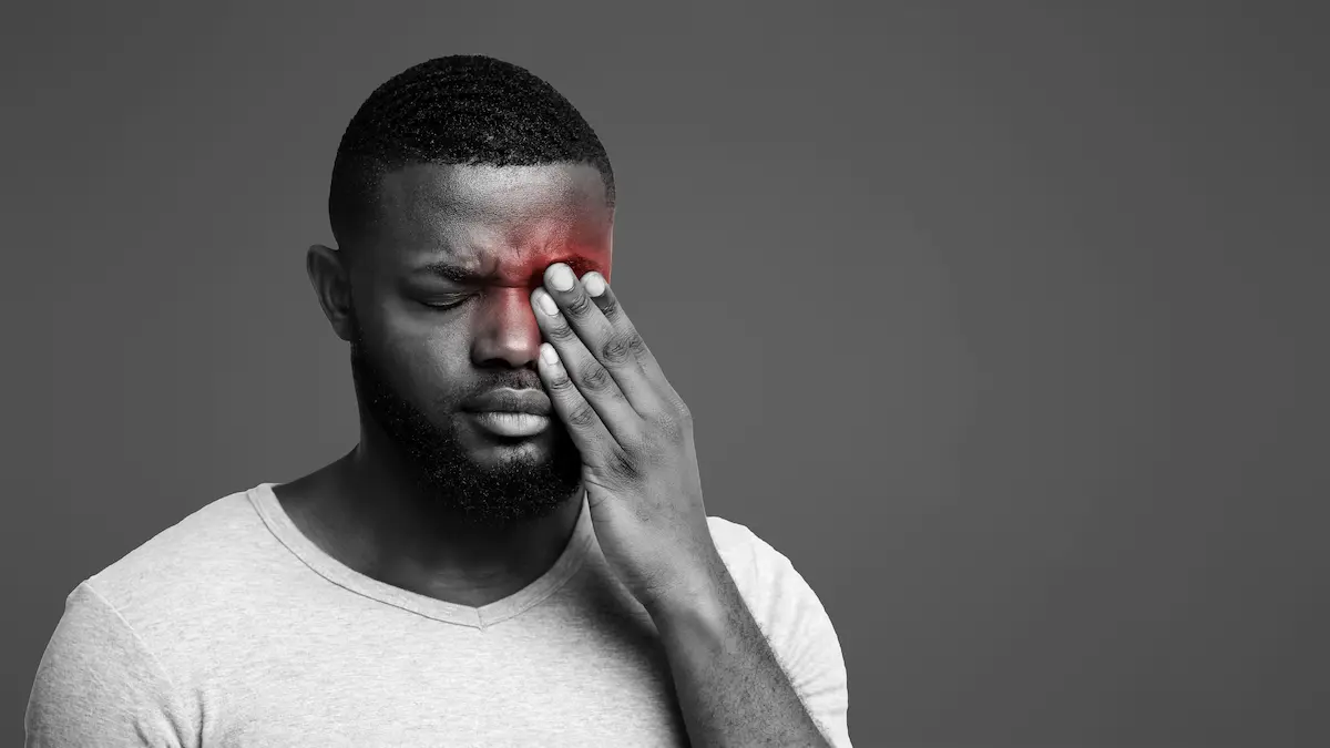 Homem negro, de olhos fechados, pressiona um dos olhos com a mão enquanto a região ao redor aparece destacada em vermelho, ilustrando uma situação relacionada a primeiros socorros oculares.