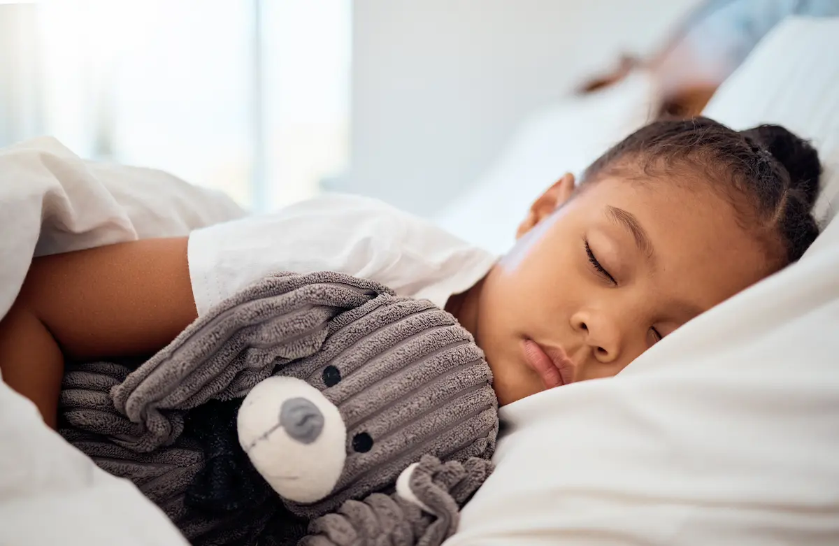 Menina negra dorme abraçada a um bicho de pelúcia na cama, em imagem relacionada ao que acontece enquanto dormimos.